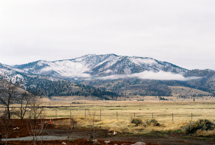 [Some short mountains have visible snow on the ground amid the green trees. There are low clouds partially covering lower levels of this mountain (but not the tops of it). The fields below the mountain appear to be dried grass.]